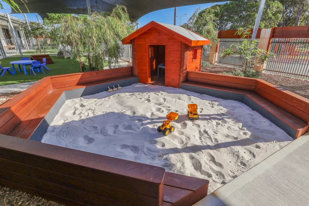 Outdoor sandpit area at Buttercups Perth Childcare featuring a wooden cubby house, toy trucks, and shaded seating, providing a fun and engaging space for creative play.