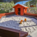 Outdoor sandpit area at Buttercups Perth Childcare featuring a wooden cubby house, toy trucks, and shaded seating, providing a fun and engaging space for creative play.