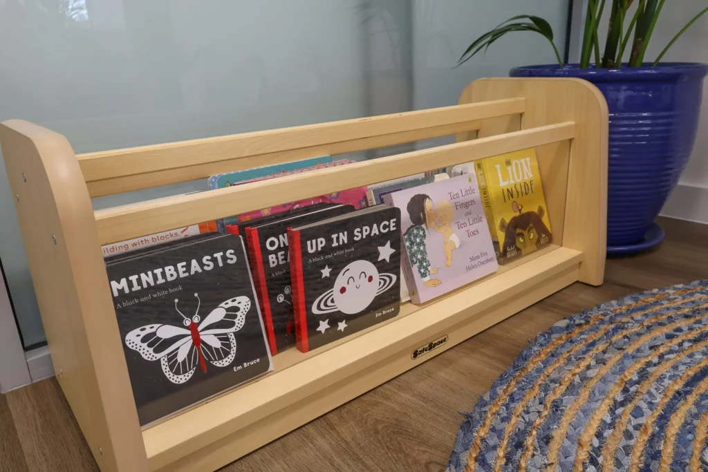 A wooden bookshelf at Buttercups Perth Childcare, filled with children's books like "Minibeasts" and "Up in Space," beside a blue plant pot and woven rug.