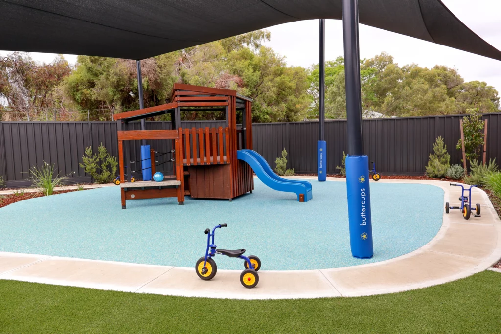 Outdoor play area at Buttercups Perth Childcare featuring a shaded playground with a wooden fort and slide, soft fall flooring, and tricycles, encouraging active play, social interaction, and physical development in a safe environment.