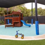 Outdoor play area at Buttercups Perth Childcare featuring a shaded playground with a wooden fort and slide, soft fall flooring, and tricycles, encouraging active play, social interaction, and physical development in a safe environment.