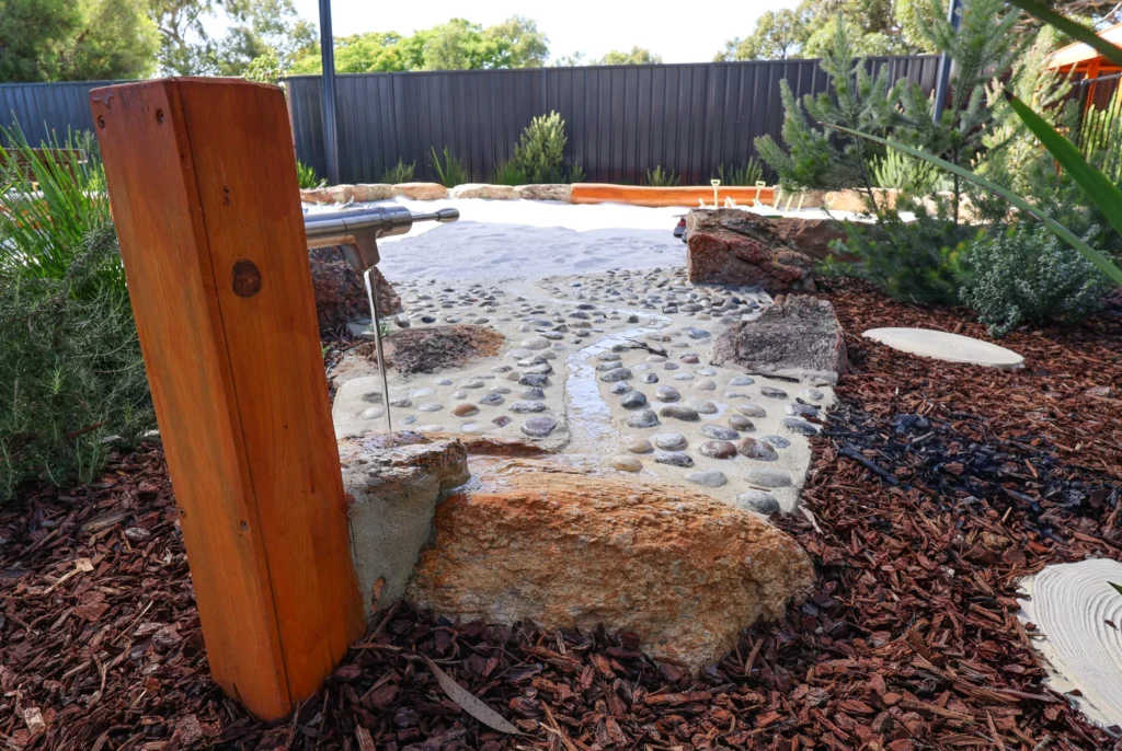 Nature-inspired water play area at Buttercups Perth Childcare, featuring a flowing water pump, smooth river stones, and a sandy play zone, designed to encourage sensory exploration and imaginative outdoor learning in a natural setting.