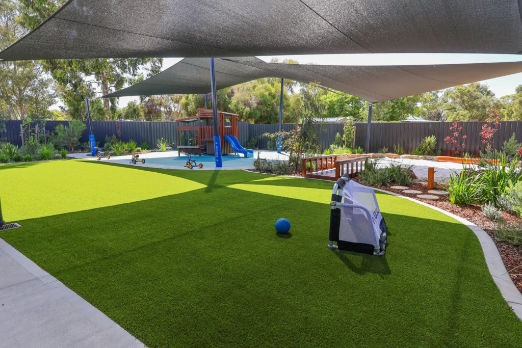 Expansive outdoor play area at Buttercups Perth Childcare, featuring shaded spaces, lush greenery, a wooden play structure with slides, and a variety of play equipment designed to encourage active exploration and imaginative play in a safe and engaging environment.