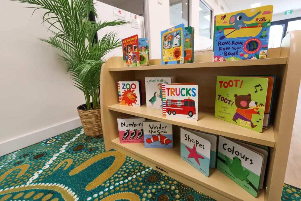 Cosy reading nook at Buttercups Perth Childcare, featuring a beautifully arranged bookshelf filled with colourful children's books, a vibrant cultural rug, and lush greenery, creating an inviting space for early literacy and exploration.