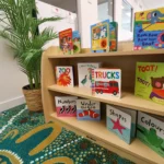 Cosy reading nook at Buttercups Perth Childcare, featuring a beautifully arranged bookshelf filled with colourful children's books, a vibrant cultural rug, and lush greenery, creating an inviting space for early literacy and exploration.