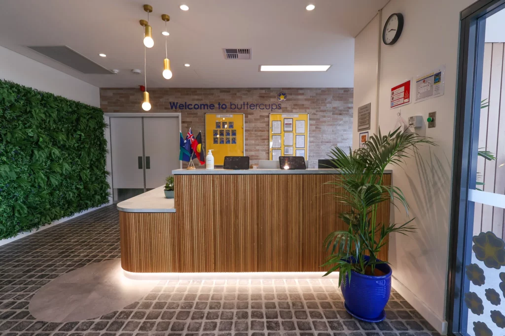 Welcoming reception area at Buttercups Perth Childcare, featuring a modern design with natural textures, greenery, and warm lighting, creating a friendly and inviting space for families and visitors.