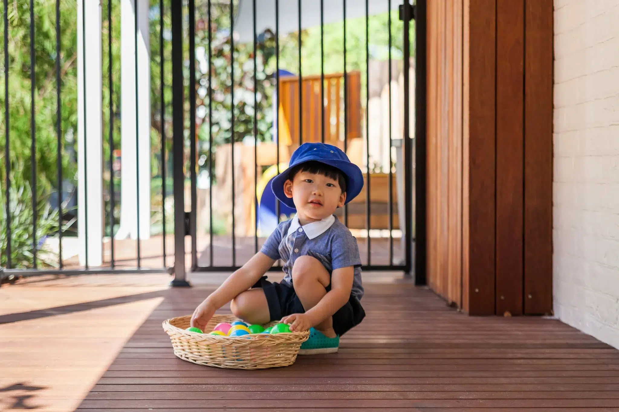 A young child at Buttercups Perth Childcare sits on a wooden deck, engaged in play with a basket of colourful toys. Wearing a blue hat and comfortable clothing, the child enjoys a moment of curiosity and exploration in a safe and nurturing learning environment.