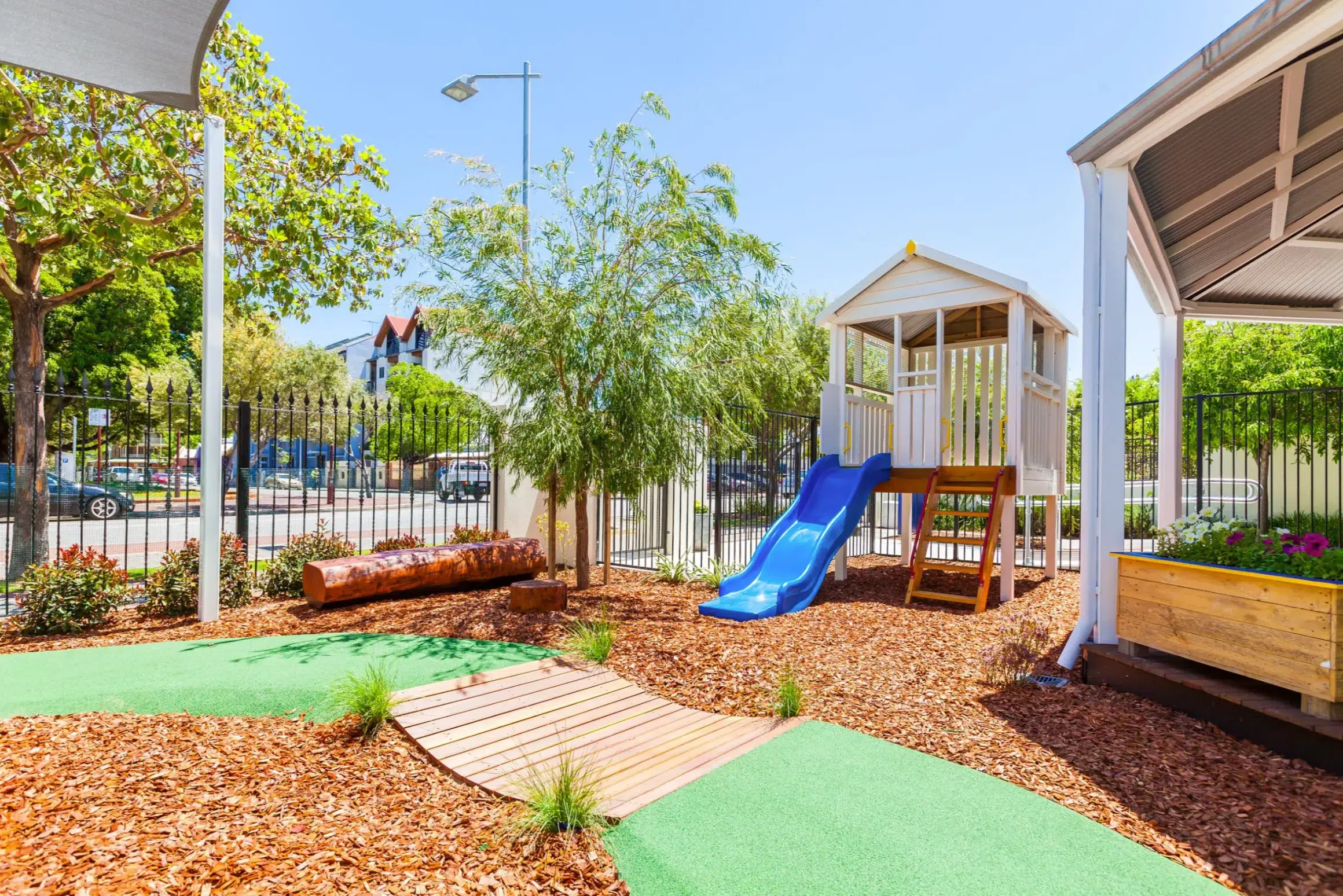 A natural outdoor play area at Buttercups Perth Childcare, featuring a wooden cubby house with a blue slide, a wooden log for seating, and a garden bed, all surrounded by greenery and shaded spaces, providing a fun and engaging environment for children.