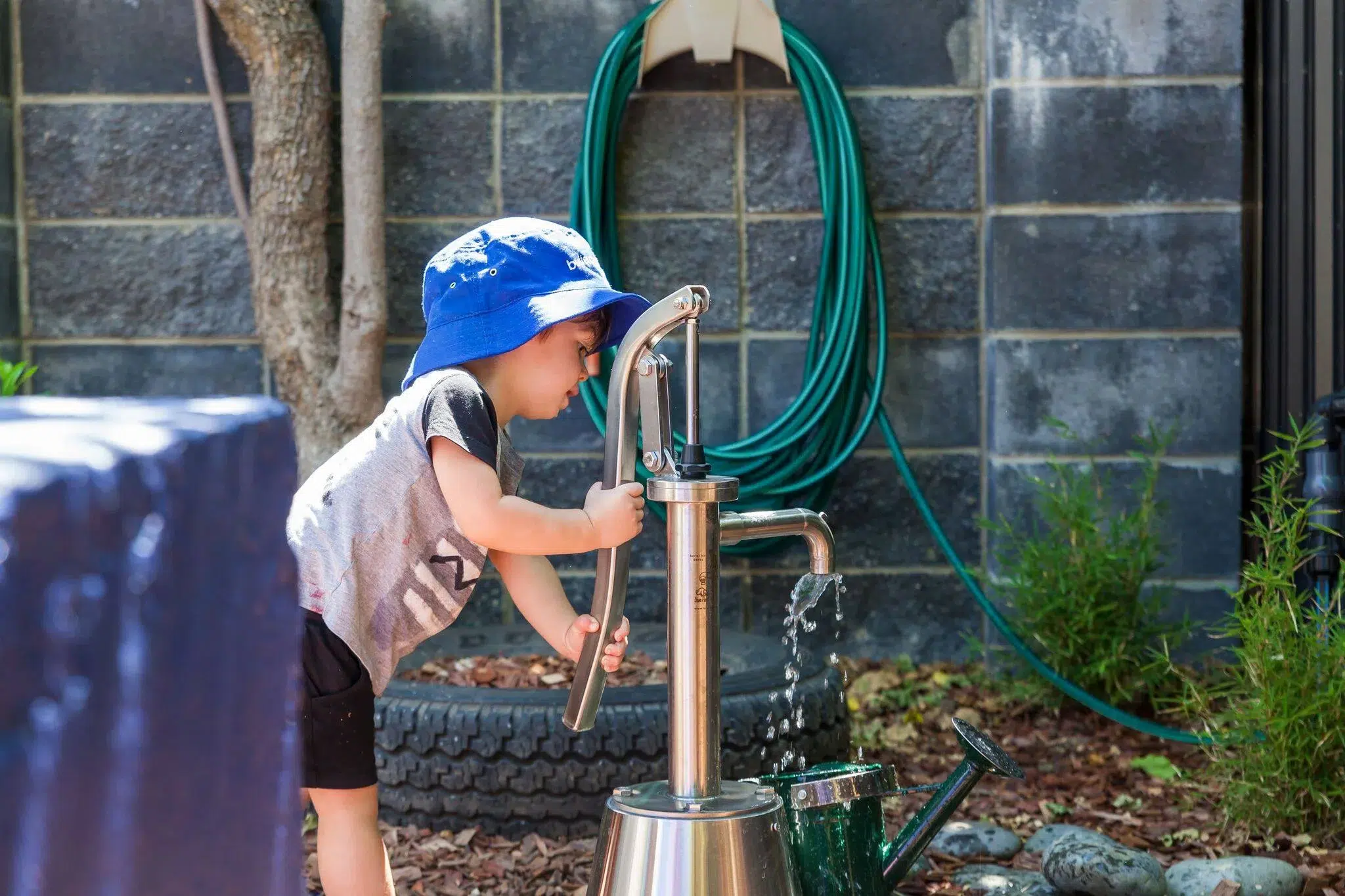 A young child in a blue hat engages with a water pump, filling a watering can at Buttercups Perth Childcare. The natural play area features a brick wall, coiled garden hose, and surrounding greenery, promoting hands-on learning and outdoor exploration.