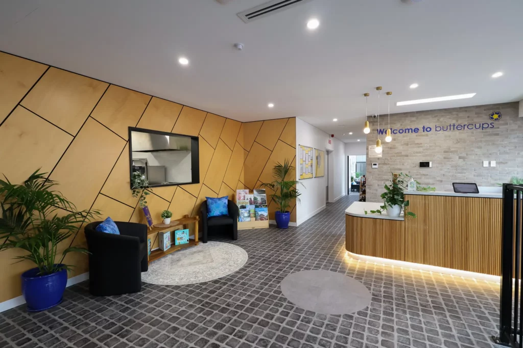 Reception area at Buttercups Perth Childcare, featuring modern wooden panels, comfortable seating, and a reception desk with a 'Welcome to Buttercups' sign. The space is bright and welcoming with plants and colourful decor, designed to create a warm atmosphere for families.