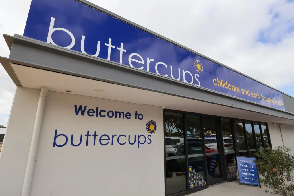 The exterior of Buttercups Perth Childcare, featuring a prominent "Welcome to Buttercups" sign, the brand logo with a yellow flower, and the words "Childcare and Early Learning Centre" on a modern building. The entrance is framed with large glass doors, and a sign promoting childcare services is placed outside.