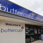 The exterior of Buttercups Perth Childcare, featuring a prominent "Welcome to Buttercups" sign, the brand logo with a yellow flower, and the words "Childcare and Early Learning Centre" on a modern building. The entrance is framed with large glass doors, and a sign promoting childcare services is placed outside.