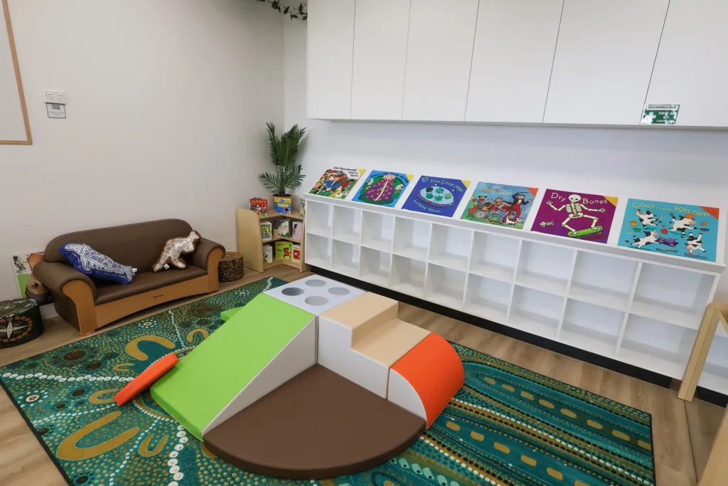 A cozy reading nook at Buttercups Perth Childcare with a soft brown sofa, colorful books neatly arranged on a low shelf, and a vibrant patterned rug. In the foreground, a multi-colored play structure with a slope and foam blocks invites exploration.