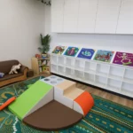 A cozy reading nook at Buttercups Perth Childcare with a soft brown sofa, colorful books neatly arranged on a low shelf, and a vibrant patterned rug. In the foreground, a multi-colored play structure with a slope and foam blocks invites exploration.