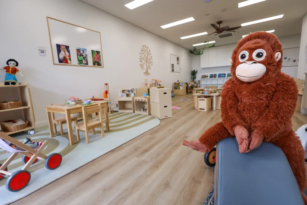 Indoor play area at Buttercups Perth Childcare featuring a wooden table with small chairs, soft rugs, and educational toys. A plush orangutan sits on the edge of a blue bench in the foreground, with playful decorations and a natural aesthetic enhancing the welcoming environment.