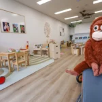 Indoor play area at Buttercups Perth Childcare featuring a wooden table with small chairs, soft rugs, and educational toys. A plush orangutan sits on the edge of a blue bench in the foreground, with playful decorations and a natural aesthetic enhancing the welcoming environment.