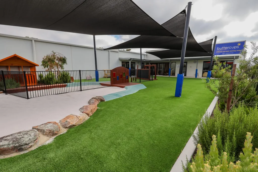 View of the outdoor play area at Buttercups Perth Childcare, featuring a large sandpit separated by rocks from the astroturf, with the Buttercups centre visible in the background, creating a safe and engaging environment for children to play and explore.