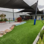 View of the outdoor play area at Buttercups Perth Childcare, featuring a large sandpit separated by rocks from the astroturf, with the Buttercups centre visible in the background, creating a safe and engaging environment for children to play and explore.