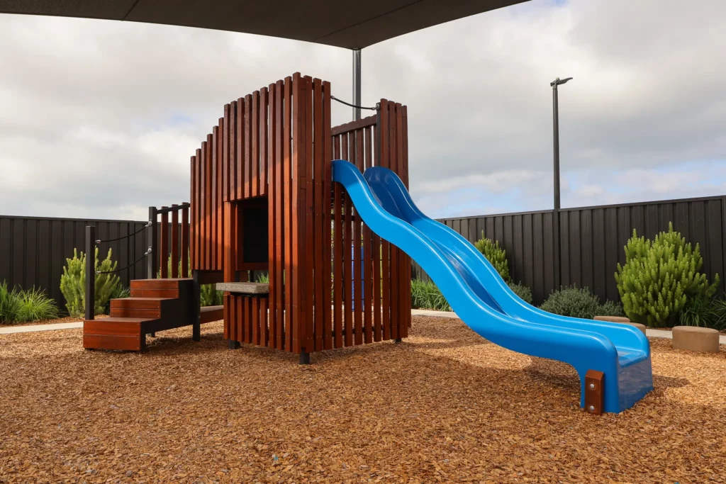 Outdoor play area at Buttercups Perth Childcare featuring a wooden frame with steps and a blue slide, all situated underneath a protective shade sail, creating a safe and exciting space for children to explore and play.