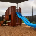 Outdoor play area at Buttercups Perth Childcare featuring a wooden frame with steps and a blue slide, all situated underneath a protective shade sail, creating a safe and exciting space for children to explore and play.