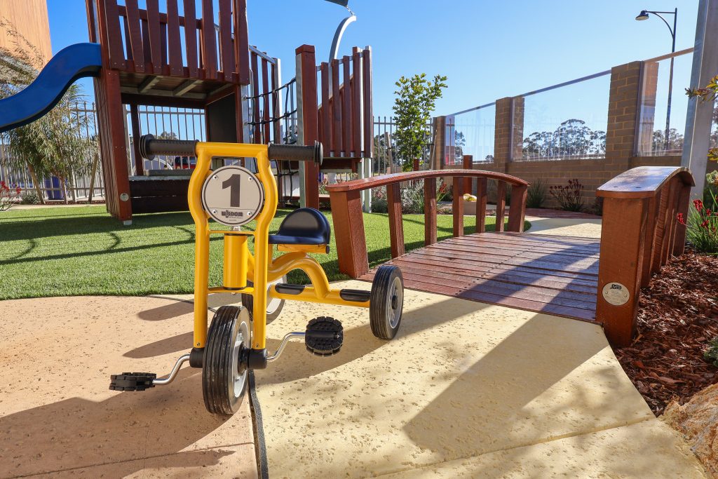 A bright yellow tricycle labelled with the number "1," parked on a clean concrete pathway in the outdoor play area at Buttercups Perth Childcare, surrounded by green artificial turf and a play fort in the background, offering children an engaging and active play experience.