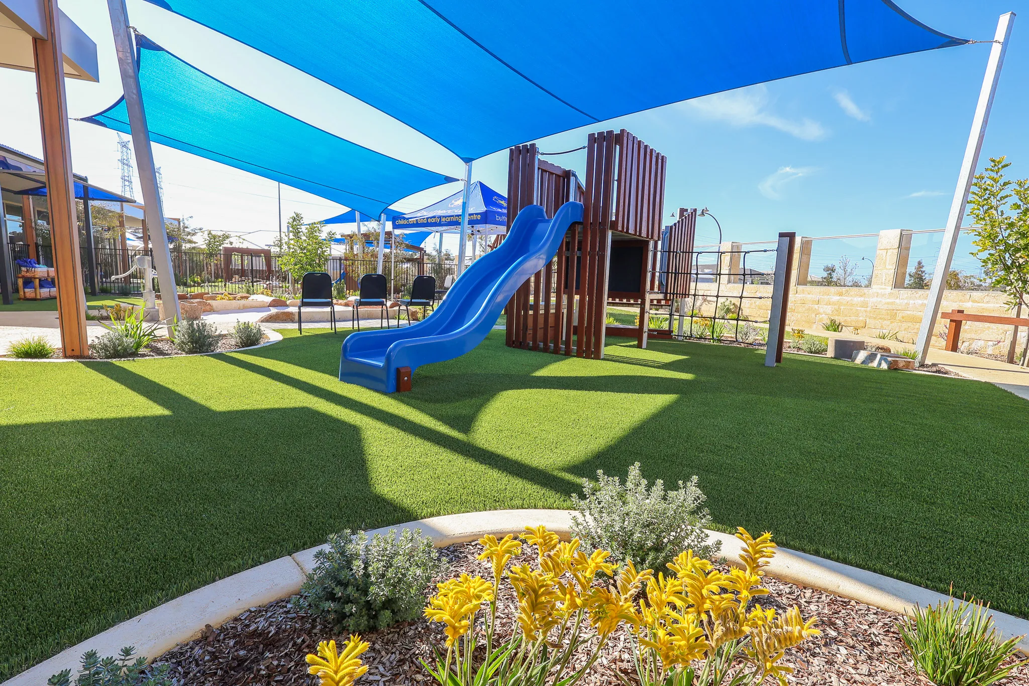 The wooden play structure with blue slide at Buttercups Perth Childcare is shaded by blue sails, offering a safe and fun environment for children.