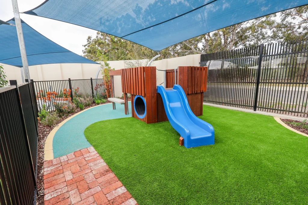 The outdoor play area at Buttercups Perth Childcare features a wooden play structure with a blue slide shaded by large blue sails.