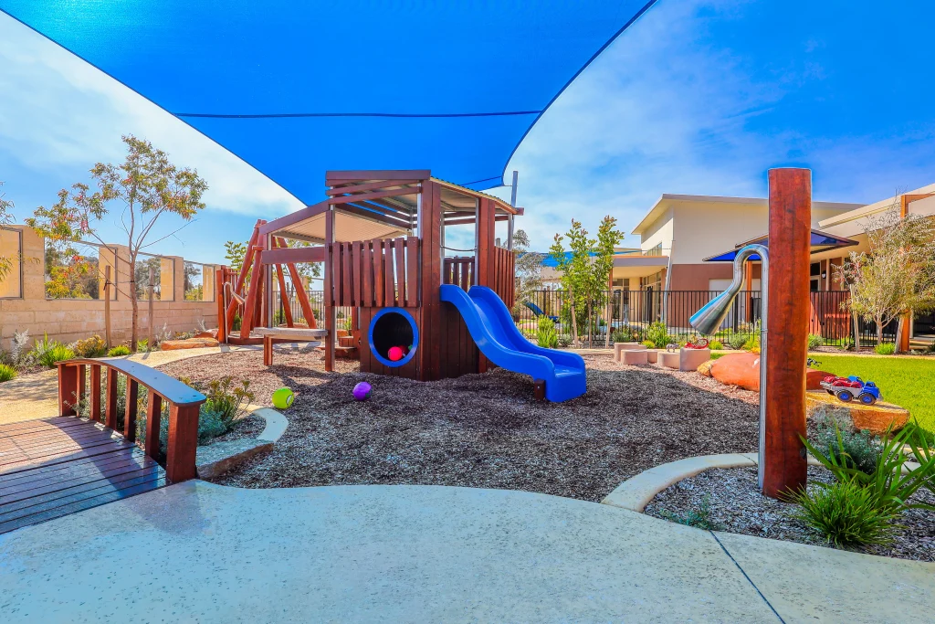The outdoor play area at Buttercups Perth Childcare features a wooden play structure with a blue slide shaded by large blue sails.