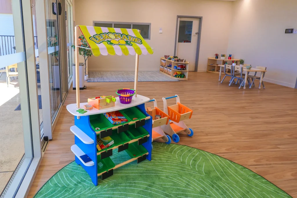Role-playing corner at Buttercups Perth Childcare with a wooden market stand stocked with toy fruits and vegetables, dress-up costumes, and a doll care station, encouraging imaginative play and creativity.