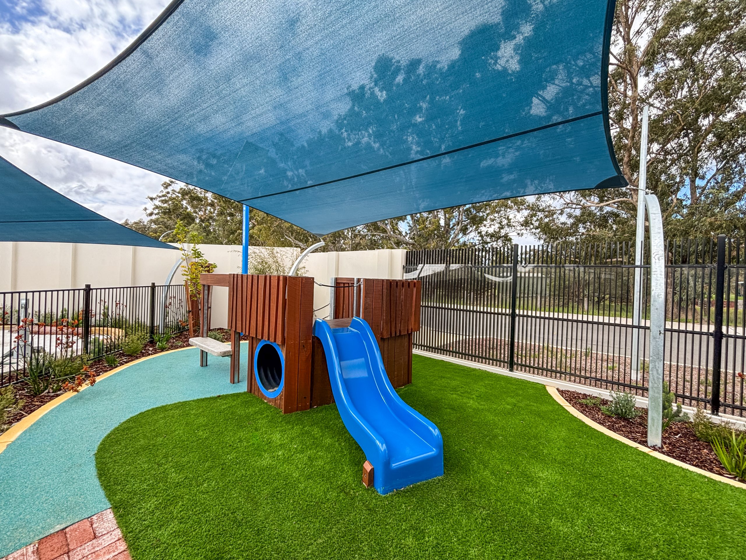 The outdoor play area at Buttercups Perth Childcare features a wooden play structure with a blue slide shaded by large blue sails.