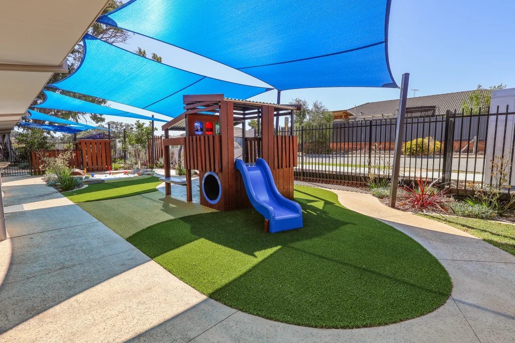 The outdoor play area at Buttercups Perth Childcare features a wooden play structure with a blue slide shaded by large blue sails.