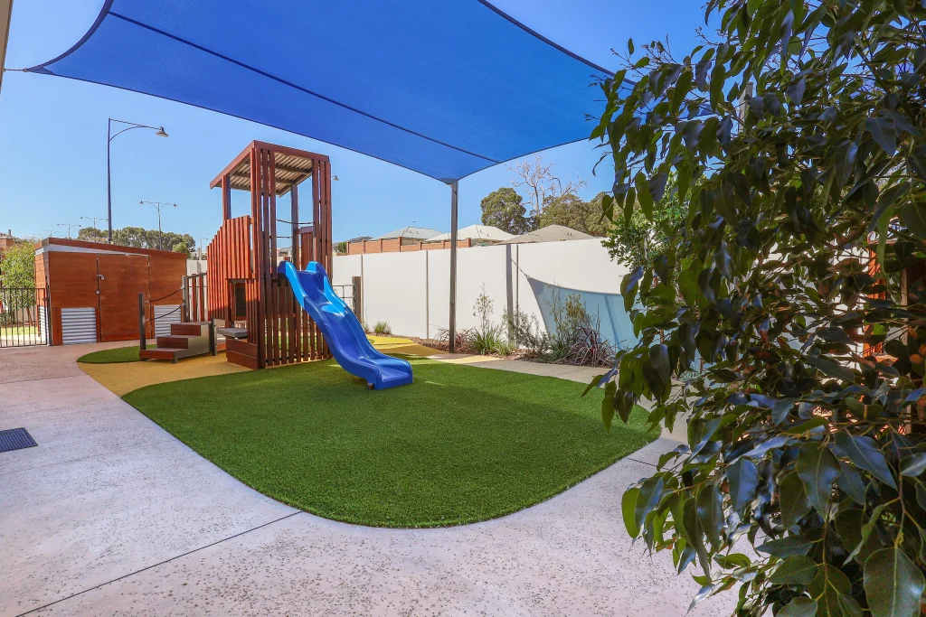 The wooden play structure with blue slide at Buttercups Perth Childcare is shaded by blue sails, offering a safe and fun environment for children.