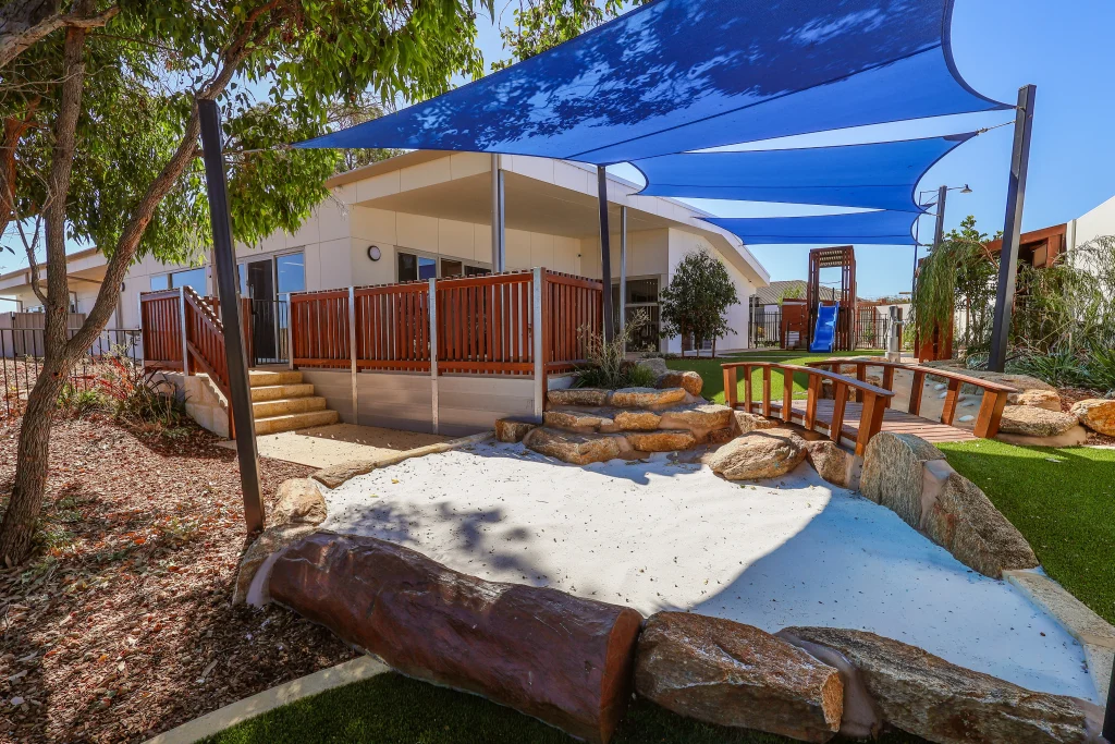 Outdoor play area at Buttercups Perth Childcare featuring a wooden frame with steps and a blue slide, all situated underneath a protective shade sail, creating a safe and exciting space for children to explore and play.