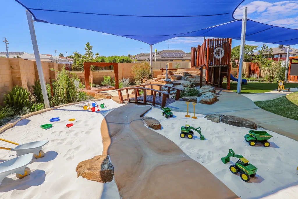 The outdoor play area at Buttercups Childcare and Early Learning Centre Lakelands features a large sandpit with a wooden bridge, rocks, and construction toys for hands-on exploration. Blue shade sails provide sun protection while children engage in sensory and imaginative play.