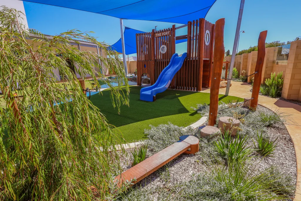 The outdoor play space at Buttercups Childcare and Early Learning Centre Lakelands includes a wooden fort with a slide, surrounded by artificial grass and natural landscaping. Balancing logs and climbing elements encourage active play and physical development under blue shade sails.