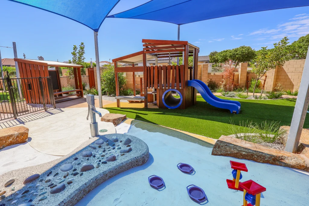 The outdoor learning environment at Buttercups Childcare and Early Learning Centre Lakelands features a water play area with a hand pump, textured surfaces, and flowing channels. This interactive space supports sensory exploration and cooperative play in a shaded setting.