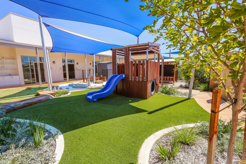 The landscaped outdoor play area at Buttercups Childcare and Early Learning Centre Lakelands includes a wooden climbing structure with a slide, surrounded by artificial grass and garden beds. Blue shade sails and natural elements create a safe and engaging environment for active play.
