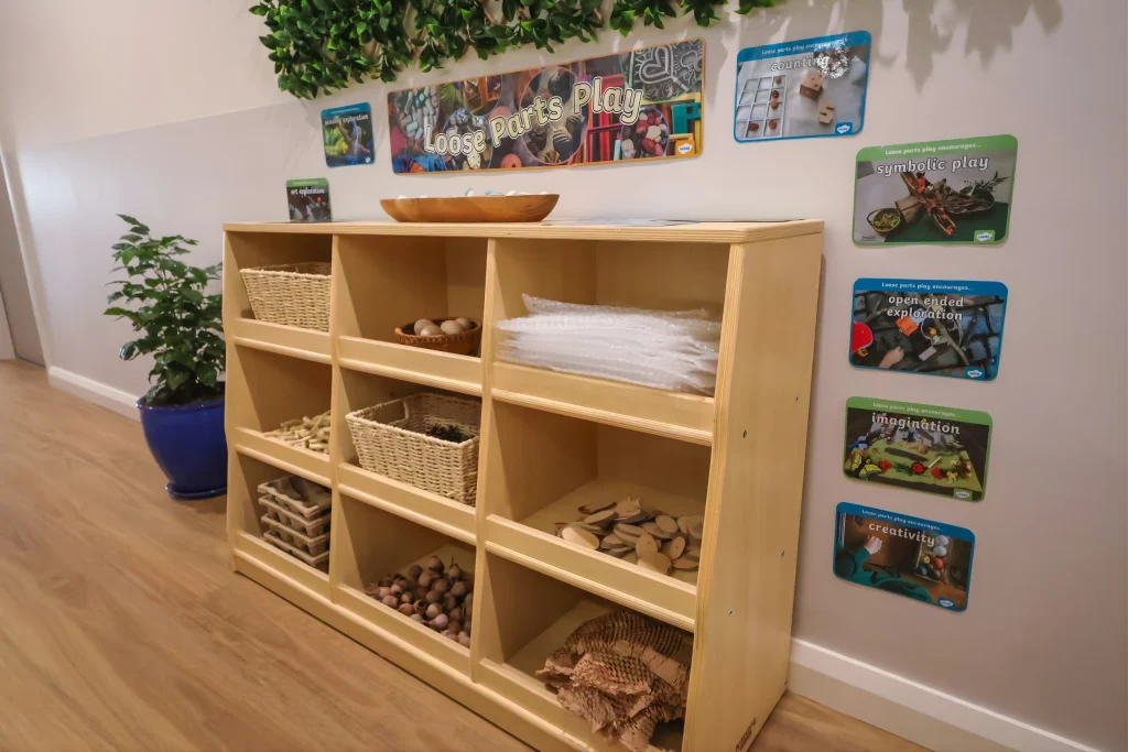 A low wooden shelving unit at Buttercups Childcare and Early Learning Centre Lakelands displays natural loose parts such as wooden discs, seed pods, and baskets. Educational signage highlights creativity, symbolic play, and open-ended exploration.