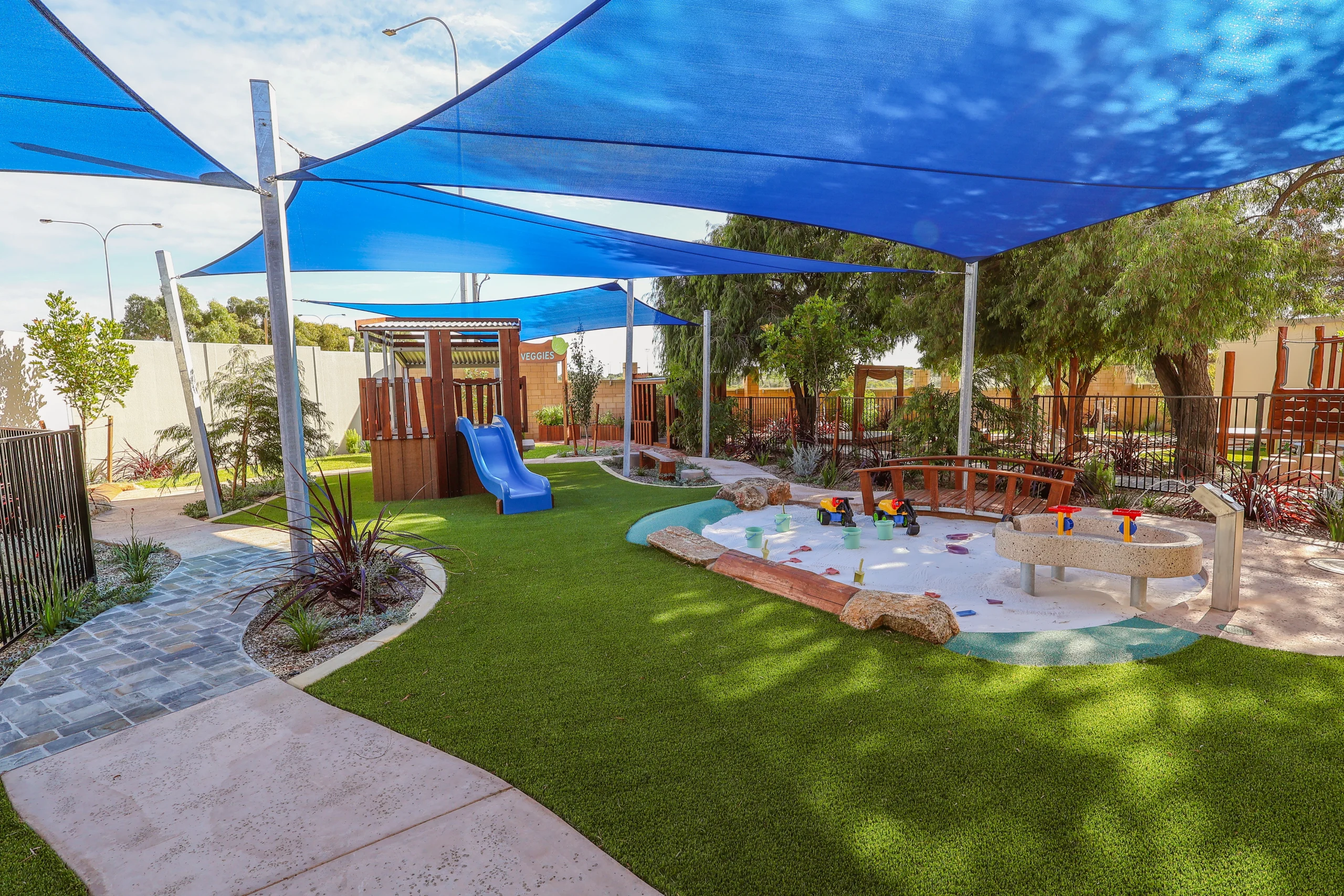 A landscaped outdoor play area at Buttercups Childcare and Early Learning Centre Lakelands features artificial grass, winding pathways, a sand and water play zone, and climbing structures. Blue shade sails provide sun protection across the space.