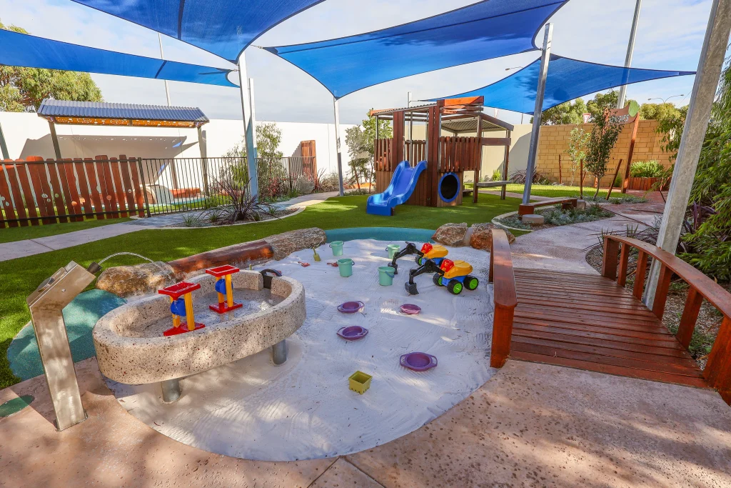 A wide view of the outdoor play environment at Buttercups Childcare and Early Learning Centre Lakelands shows a large sandpit with water play features, a wooden bridge, and surrounding play equipment. Blue shade sails and artificial grass create a safe, engaging space.