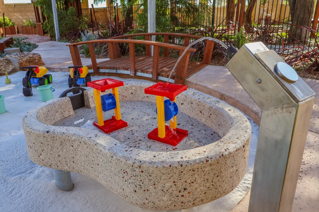 A close-up of the outdoor water play station at Buttercups Childcare and Early Learning Centre Secret Harbour shows a hand pump flowing into a concrete basin with colourful spinning elements. Sand toys and a small wooden bridge surround the area, supporting sensory and exploratory play.