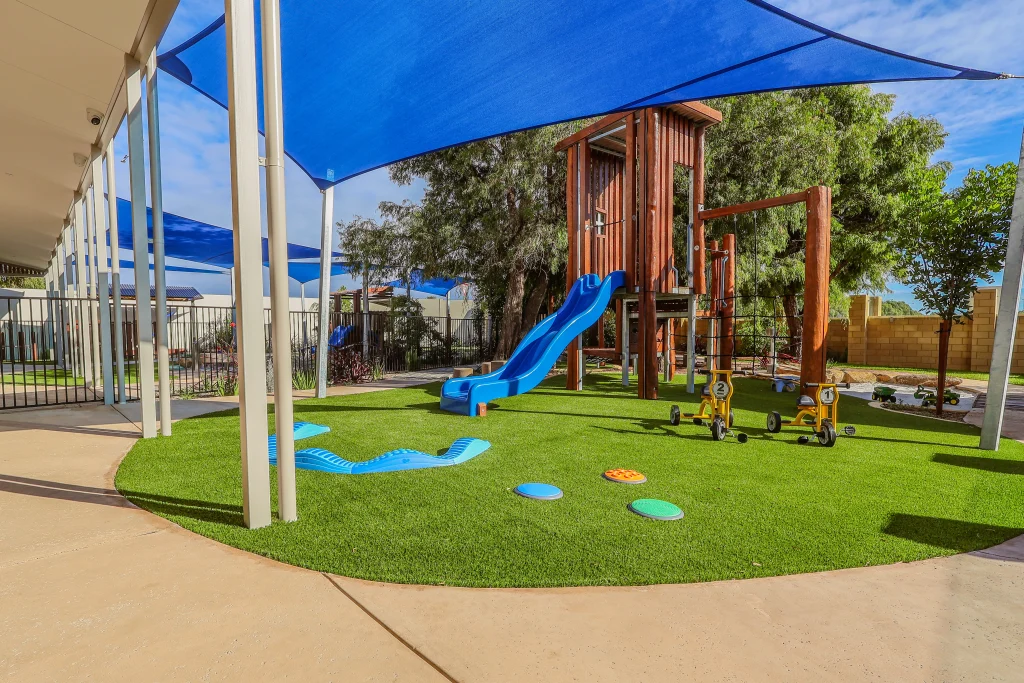 The outdoor play area at Buttercups Childcare and Early Learning Centre Lakelands features a wooden climbing structure with a blue slide, set on artificial grass. Blue shade sails provide sun protection, with tricycles and balance elements encouraging active play.
