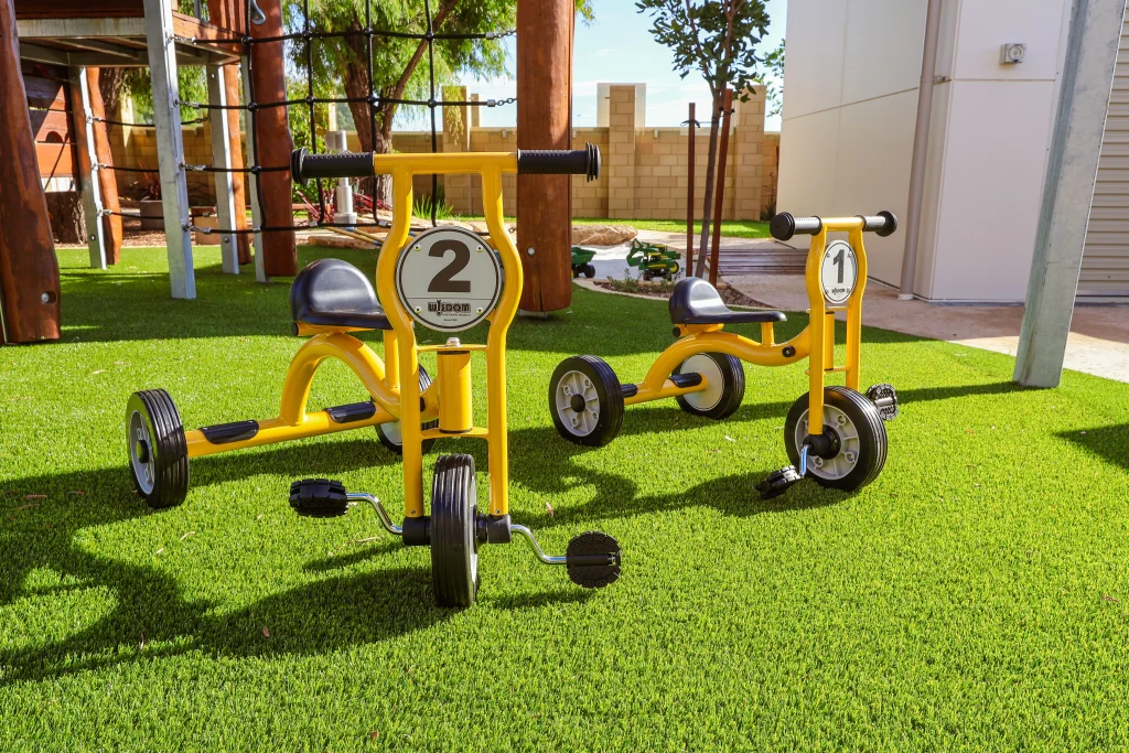 A pair of yellow tricycles sit on artificial grass in the outdoor play space at Buttercups Childcare and Early Learning Centre Lakelands. Positioned near a climbing structure, they support gross motor development and active exploration.