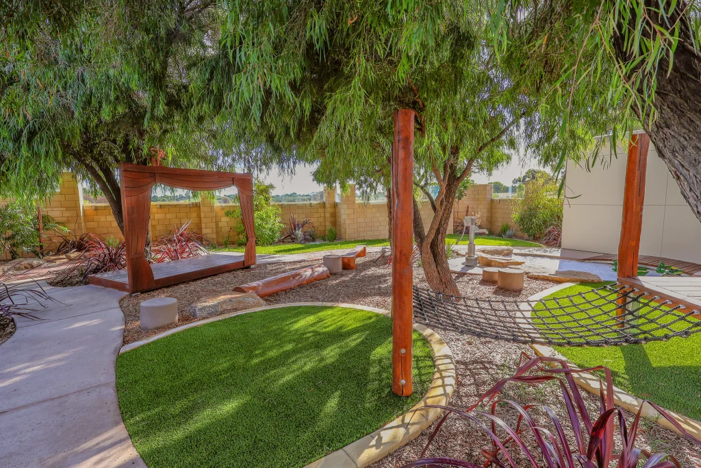 A nature-inspired outdoor play space at Buttercups Childcare and Early Learning Centre Secret Harbour features mature trees, a climbing net leading to a wooden play fort, and a timber “theatre” area with logs and stones for seating. A water play feature can be seen in the background, with soft landscaping creating a calm environment for imaginative and physical play.